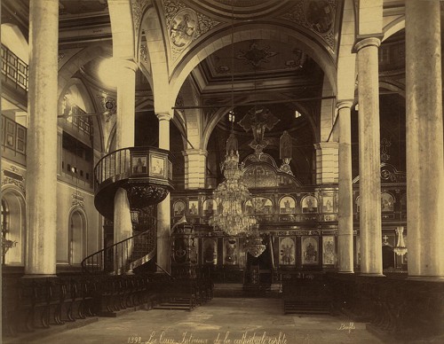 Interior of a Coptic cathedral with seats, iconostasis, icons, chandelier, and a spiral staircase leading to pulpit.